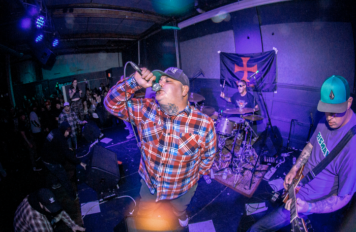 A live band performance with a vocalist singing into a microphone, wearing a plaid shirt and a cap. In the background, a drummer plays, and a guitarist is visible on the right. The venue has an energetic audience and a backdrop featuring a flag.