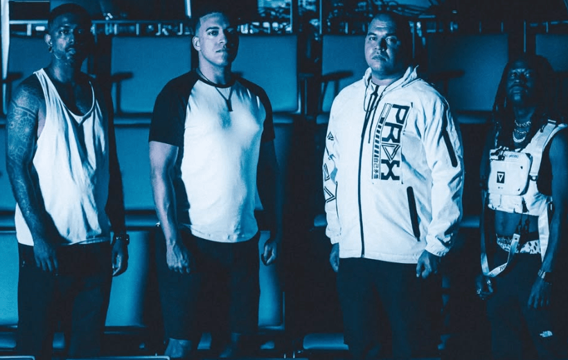 Four men standing confidently in a moody, blue-lit environment, wearing athletic clothing and displaying a strong presence.