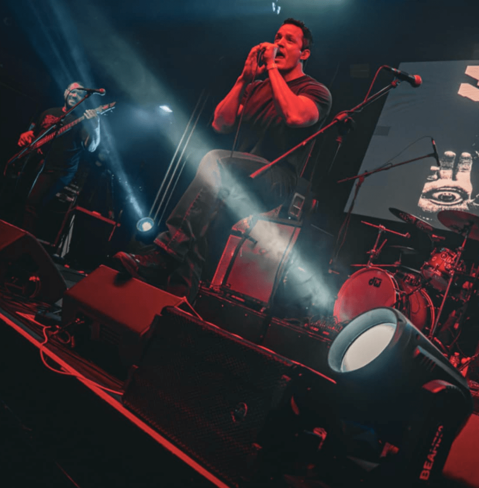 A vocalist passionately performing on stage while holding a microphone, accompanied by a bassist in the background, with stage lights and musical instruments visible.