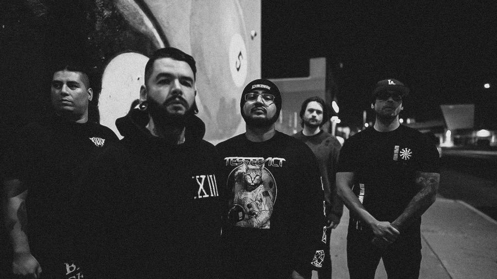 A group of six men standing together at night against a wall, wearing dark clothing, exuding a serious attitude.