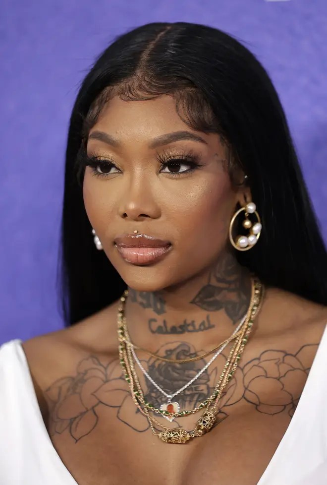 Close-up portrait of a woman with long black hair, wearing large pearl earrings and multiple layered necklaces, showcasing tattoos on her neck and chest, set against a purple background.