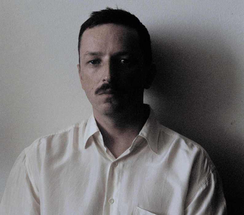 A man with a mustache wearing a white shirt, sitting in front of a light-colored wall, with a somber expression.