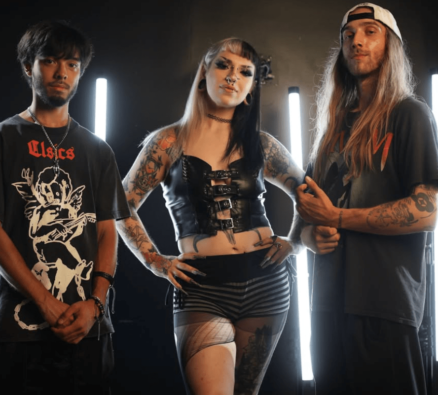 Three individuals posing together in a dimly lit setting with glowing vertical lights in the background. The person in the center has a bold fashion style featuring a black leather top and striped shorts, surrounded by two others in casual streetwear, one sporting a graphic t-shirt.