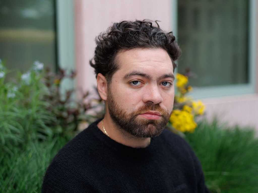 A man with curly hair and a beard is sitting outside, looking directly at the camera. He wears a black sweater, and there are colorful flowers and green plants in the background.