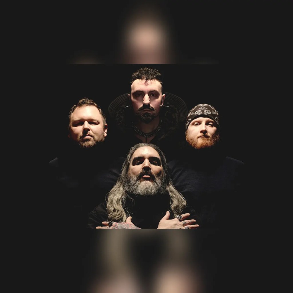 Four men posing in a dark setting, with one figure in front holding his hands in a prayer position and a long beard, while the others stand behind, giving a serious look.
