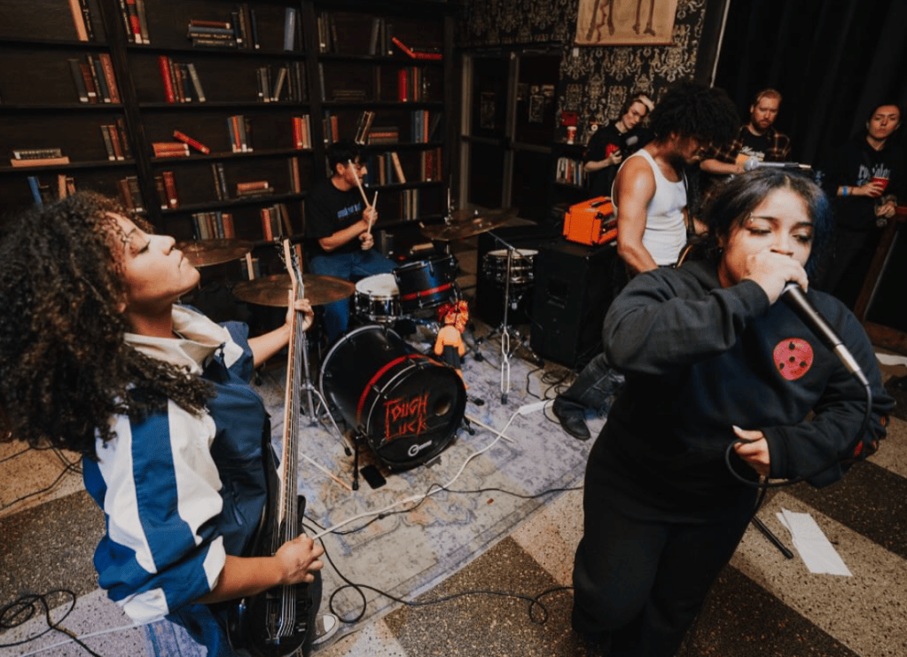 A lively band performing on stage, featuring a female vocalist holding a microphone, a woman playing bass guitar, and a male drummer in the background. The venue has a cozy atmosphere with bookshelves and audience members enjoying the performance.