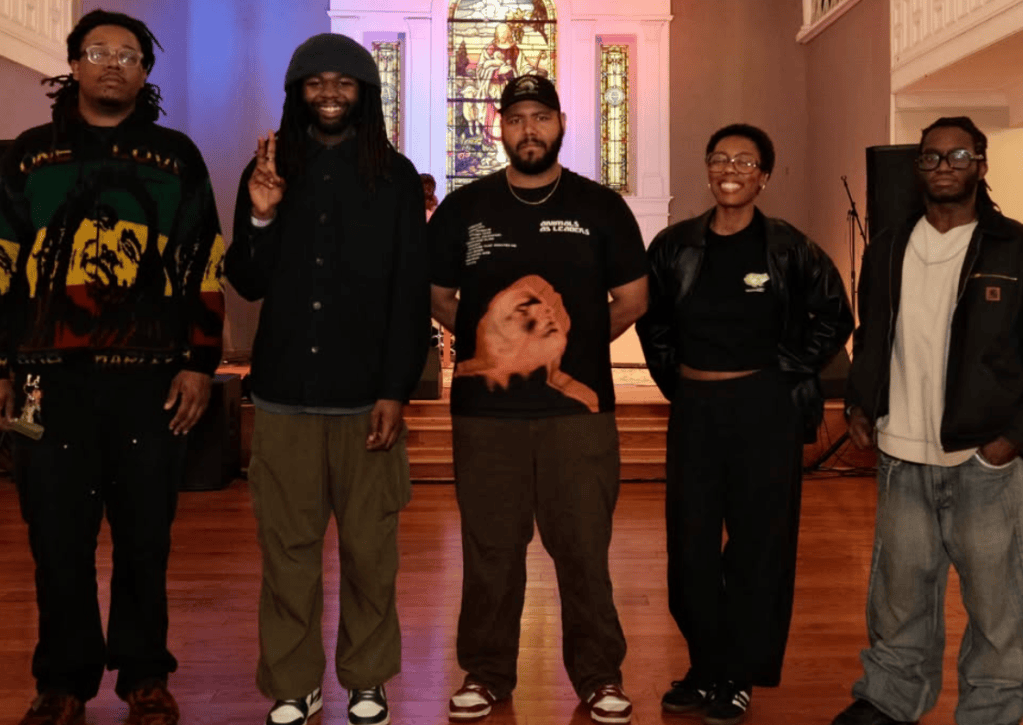 Five individuals standing together in a warmly lit indoor space, featuring a stained-glass backdrop. They are dressed in casual streetwear, with varied hairstyles and expressions.