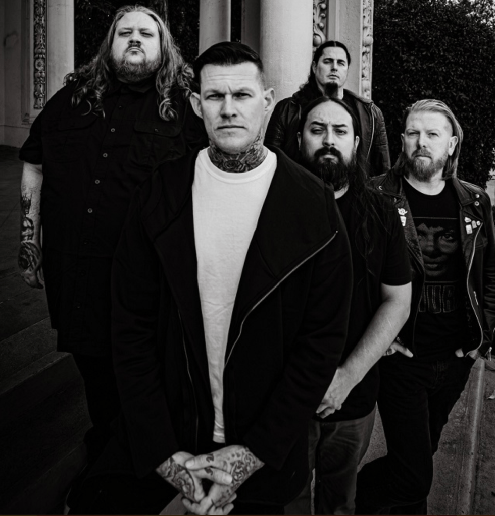 A black and white group photo of five men, each with distinct styles and tattoos, posing confidently outdoors. The front man has a serious expression and tattoos on his arms, while the others stand behind him in varying poses.