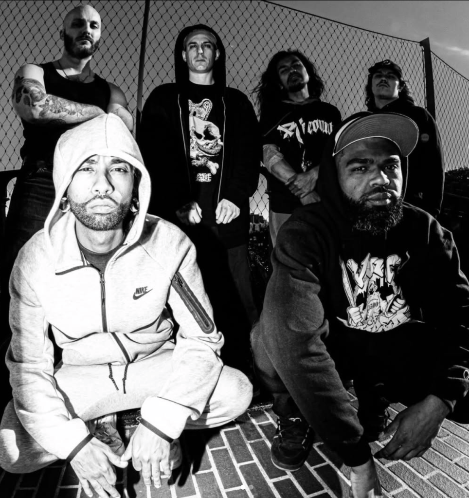 A group of six individuals posing together outdoors, with a focus on a man in a gray Nike tracksuit crouching in the front. The others stand behind him in dark clothing, against a backdrop of a chain-link fence.