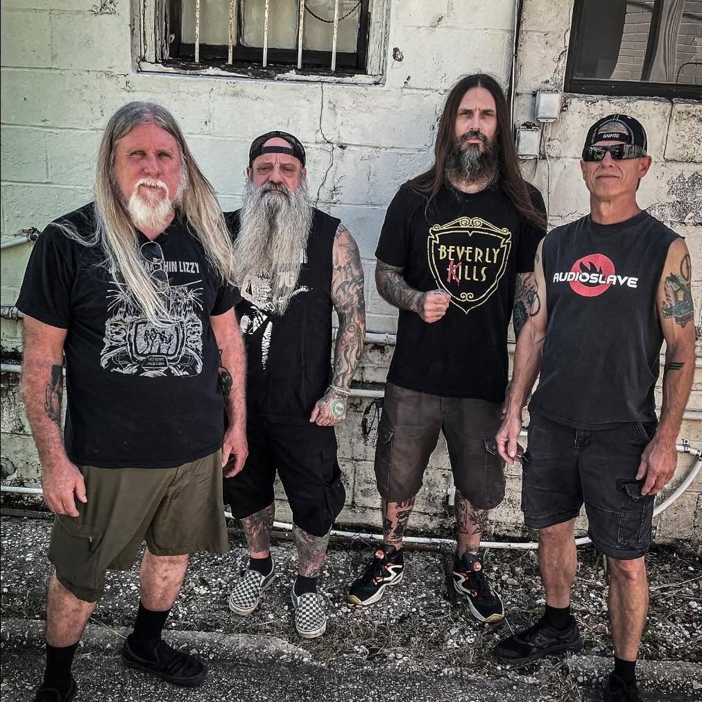 Four men with long hair and beards stand together in front of a distressed wall, wearing band t-shirts and shorts.