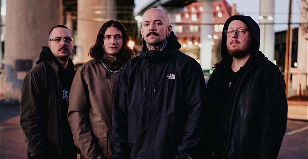 Four men standing together under an urban overpass during twilight. They are dressed in casual dark clothing and look directly at the camera.