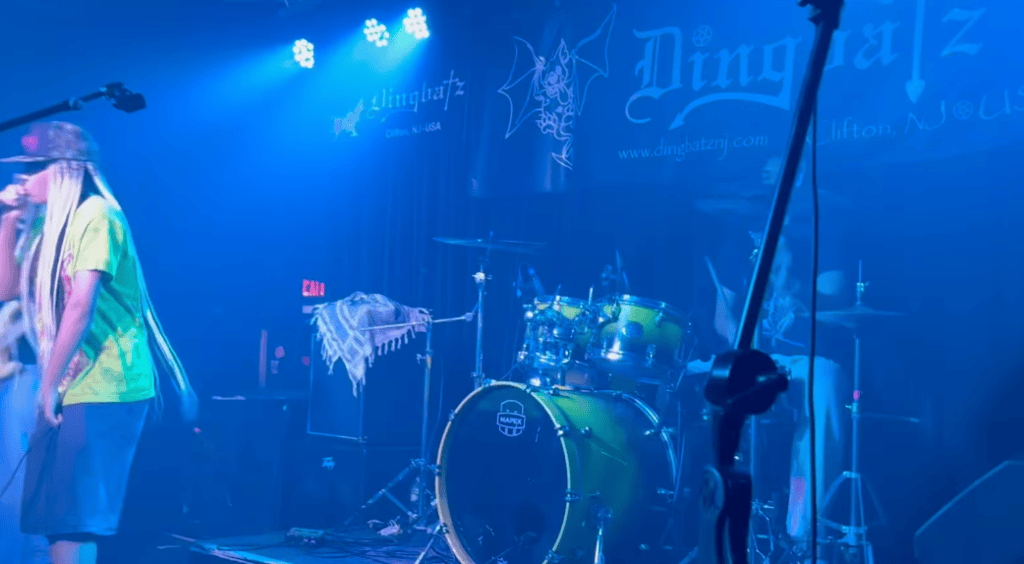 A performer singing on stage in a brightly lit venue, with a drummer and drum set visible in the background. The stage has blue lighting and a banner displaying the venue's name, Dingbatz.