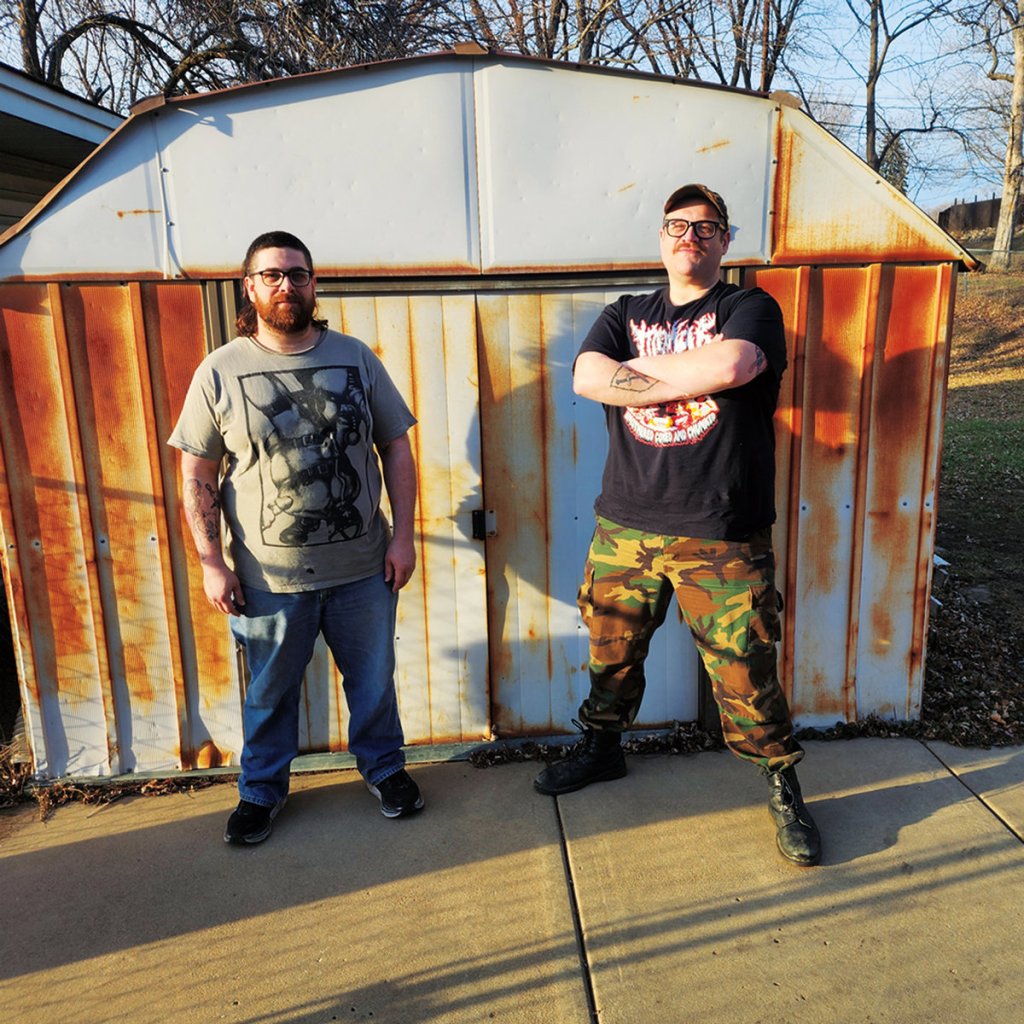 Two men standing in front of a rusty shed, one wearing a graphic t-shirt and the other in a black t-shirt and camouflage pants.