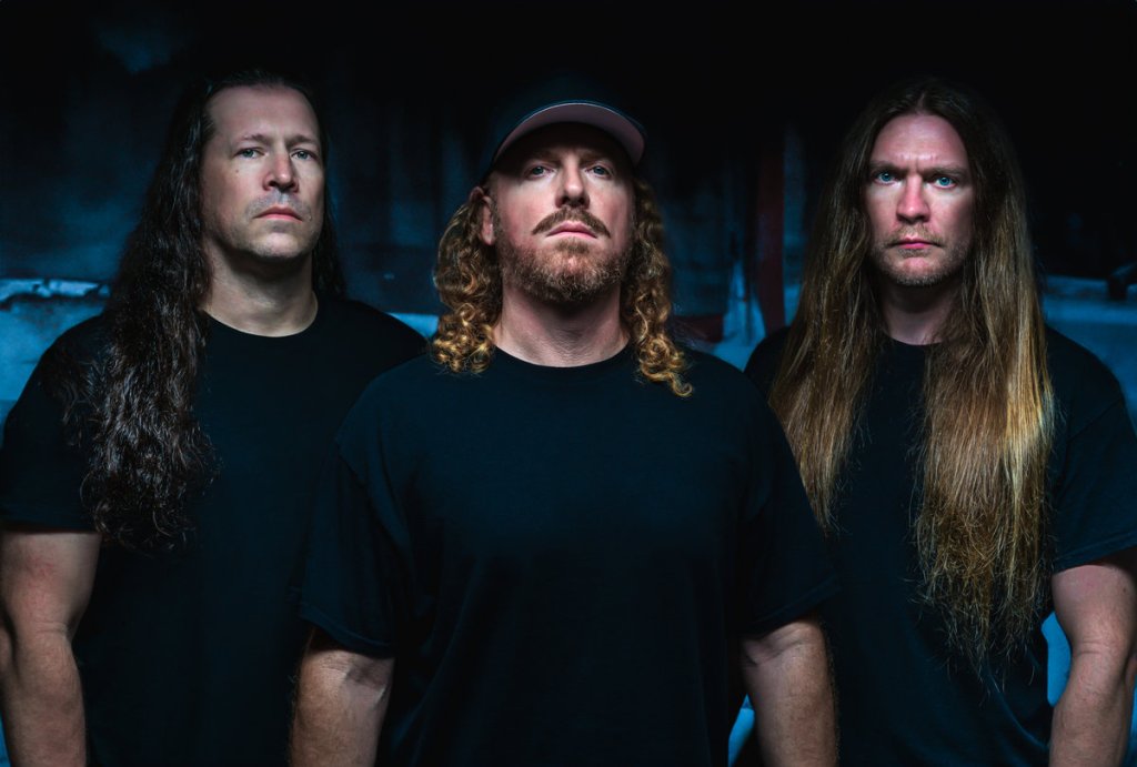 Three men standing closely together, wearing black t-shirts, with long hair; one man has curly hair and a cap, while the others have straight hair.