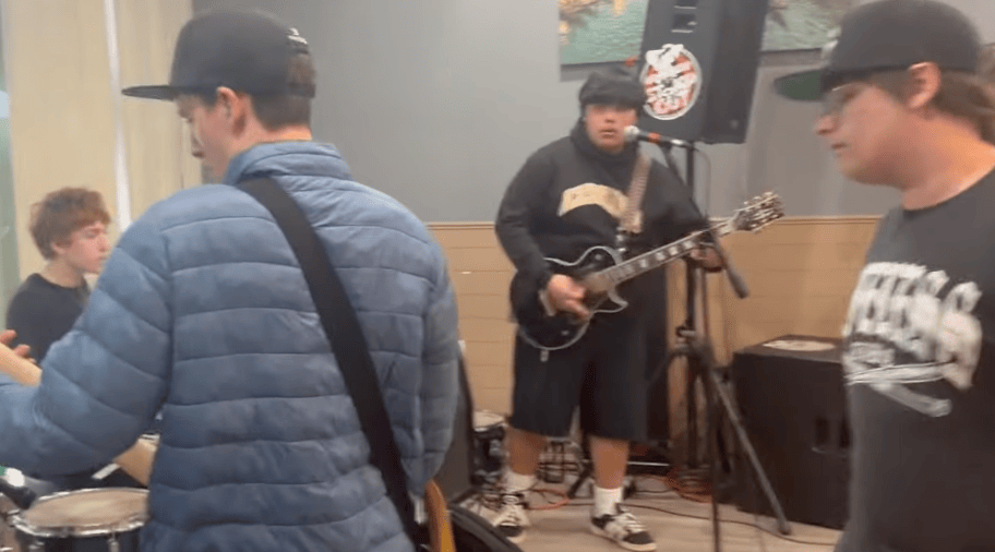 A group of young musicians performing together, with one playing a guitar while others focus on their instruments in a casual setting.