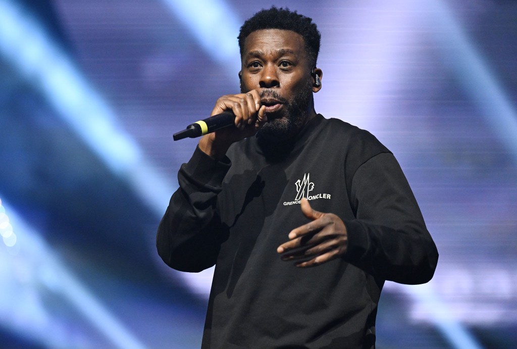 A male performer passionately rapping into a microphone on stage, wearing a black sweatshirt, with stage lights in the background.