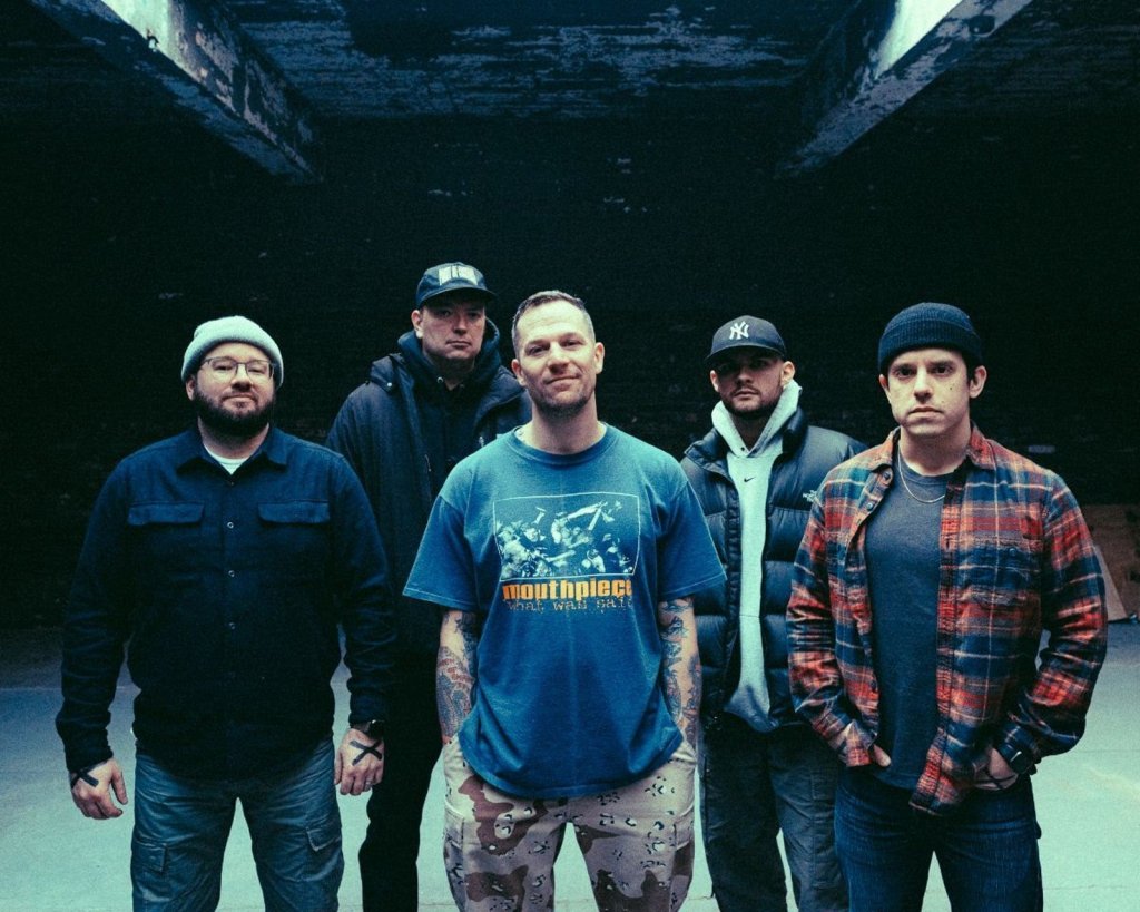 A group of five men posing together in an industrial setting, wearing casual clothing. They are standing closely, with a mix of expressions and styles, under dim lighting.