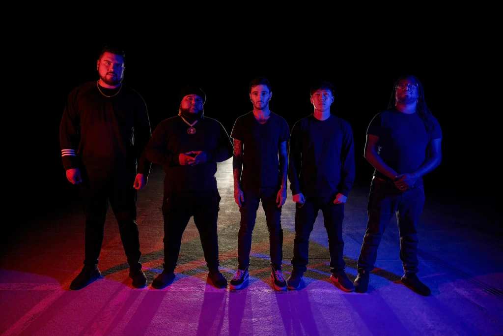 A group of five individuals standing in a dark space, illuminated by blue and purple lights, wearing dark clothing.