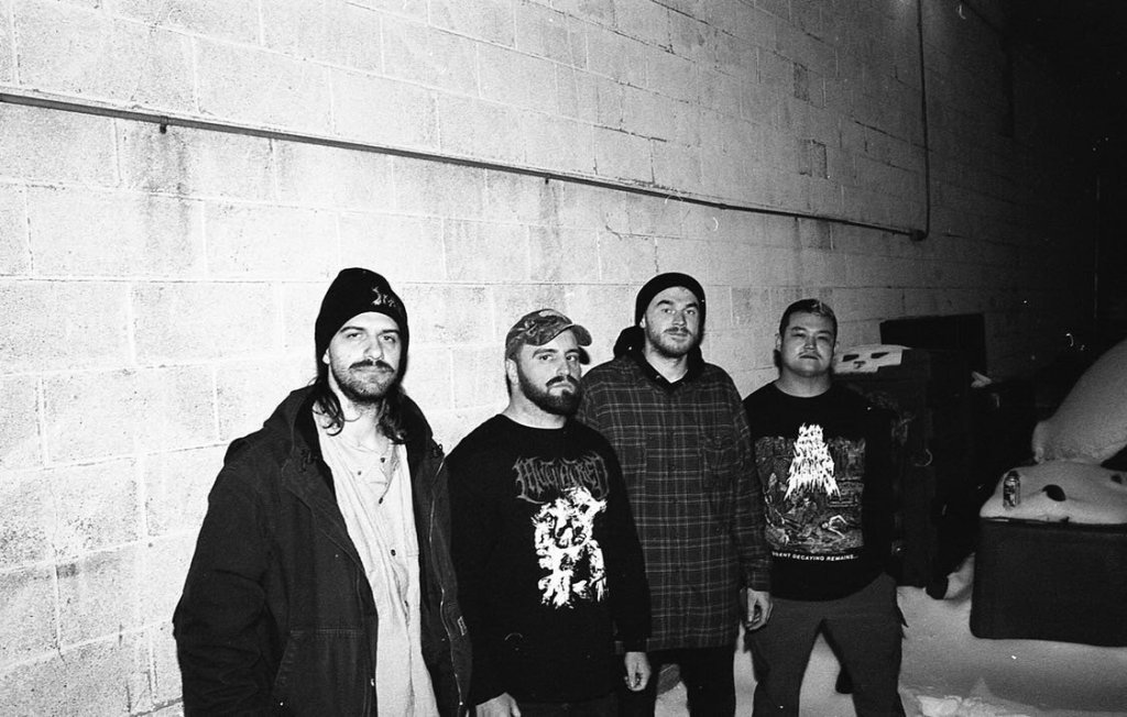 Four men standing together outside in an urban setting, dressed in casual winter clothing, against a brick wall with snow on the ground.