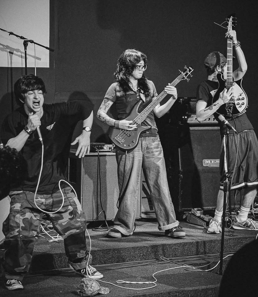 A black and white photo of a band performing on stage. The lead singer is energetically singing into a microphone, wearing a black shirt and camouflage pants. A bassist stands beside him, focused on playing, and a guitarist at the right is holding an electric guitar high. Amplifiers and musical equipment are visible in the background.