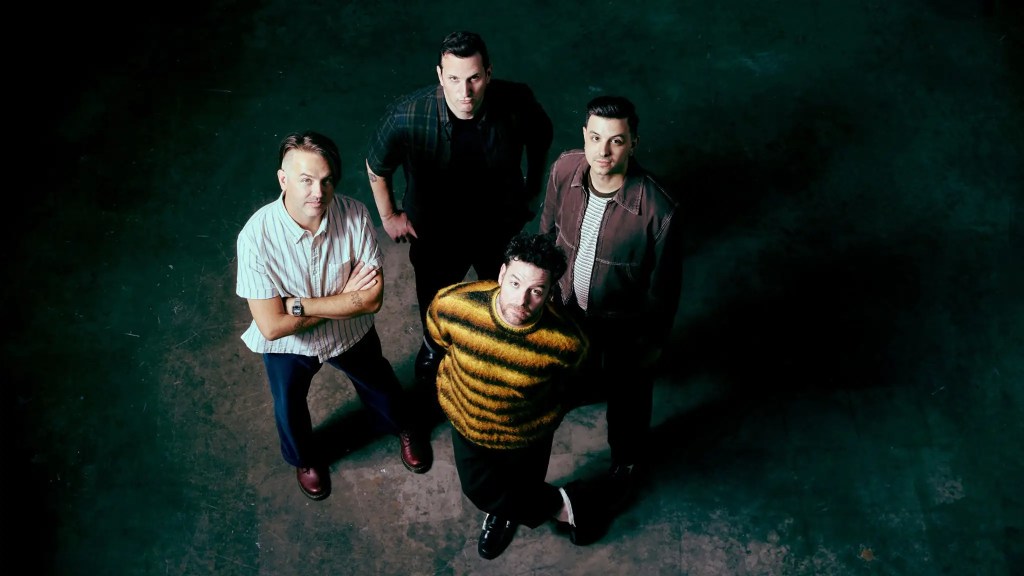A group of four men standing together, looking up at the camera, in a dimly lit space. They are dressed in casual clothing, with one wearing a striped sweater and the others in various patterned shirts.