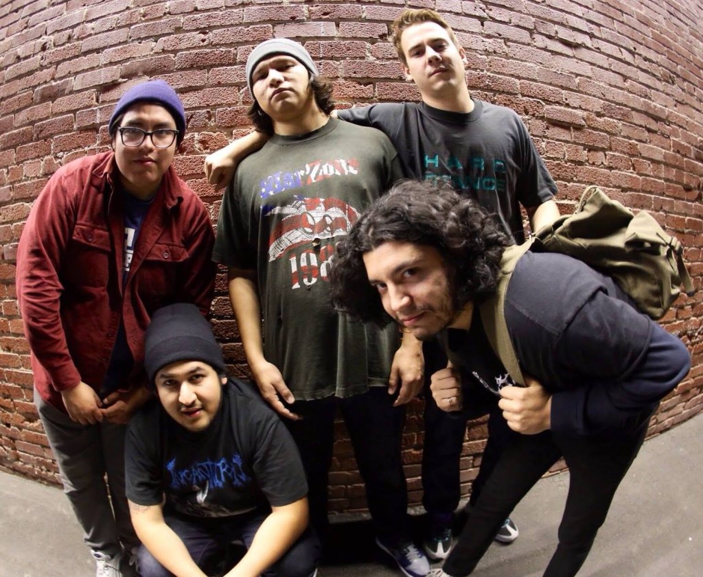 Five young men posing together in front of a brick wall, dressed in casual streetwear and band t-shirts.