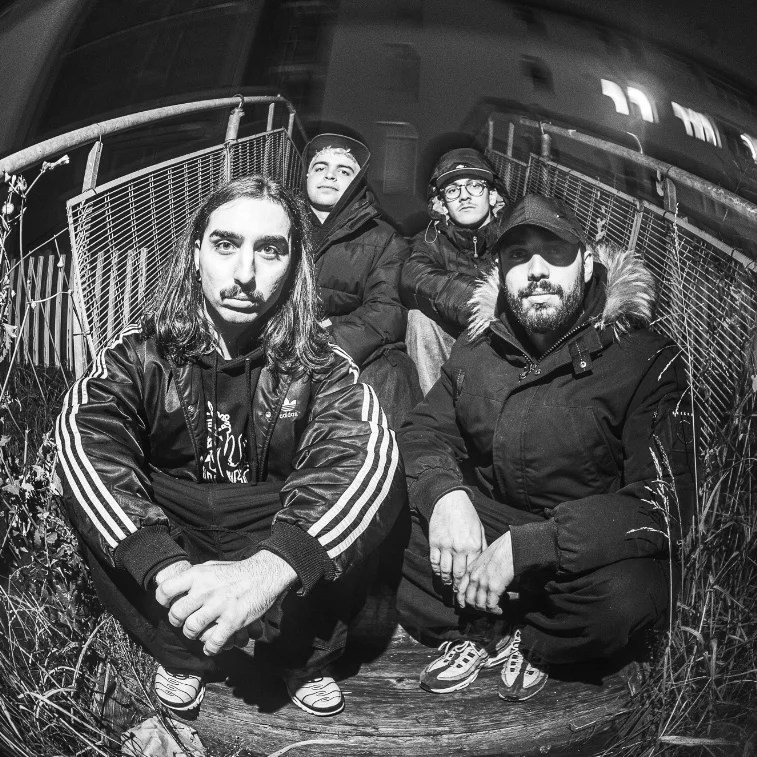 A black and white photo of four individuals sitting on a wooden surface outdoors at night, with a fence in the background. They are wearing casual winter clothing and looking directly at the camera.