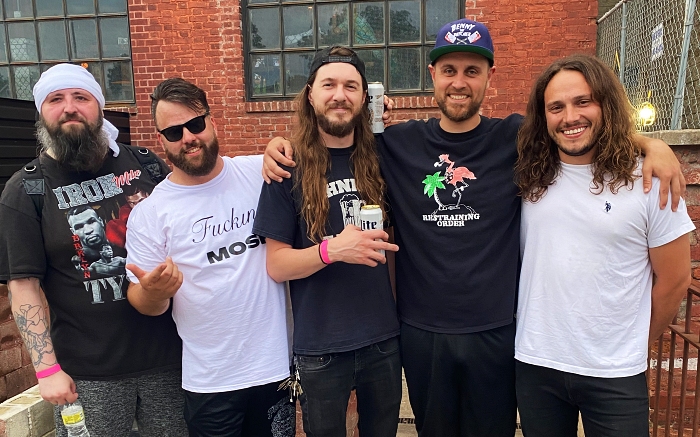 Five men posing for a photo outdoors, wearing casual clothing and smiling, with a brick wall and windows in the background.