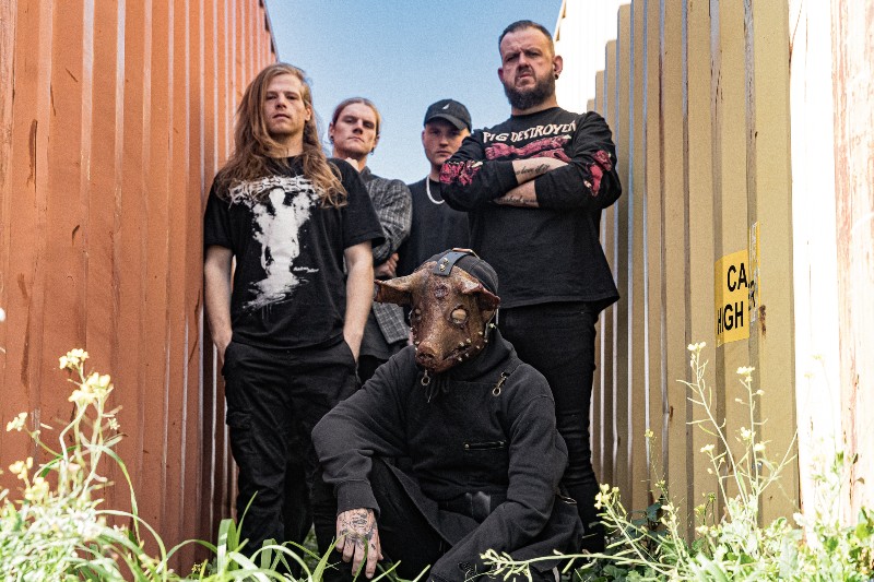 A group of five people standing in a narrow alley, with two men in the back crossing their arms and a person in the front wearing a mask resembling a cow's head.