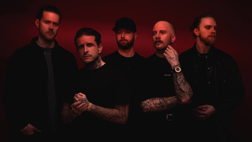 A group of five men standing in a dramatic pose against a red background, wearing mostly black clothing, showcasing various tattoos.