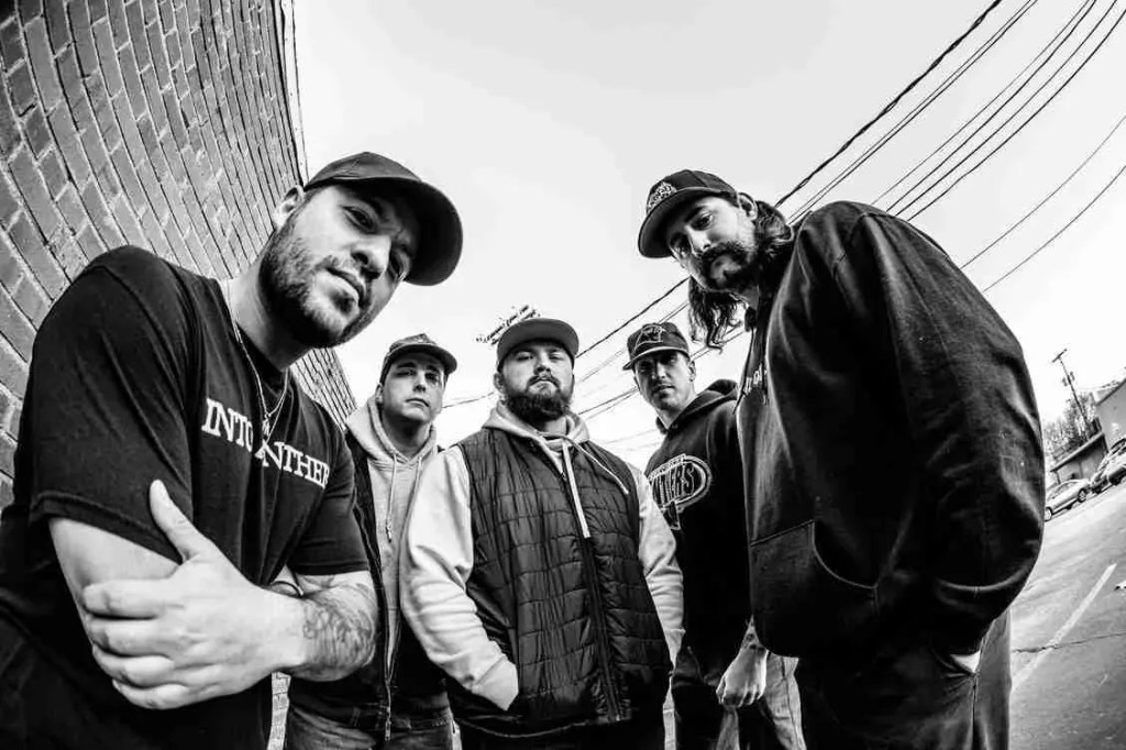 A group of five men stands together in a black and white photo, posing against a brick wall. They have a casual streetwear style, with hats and hoodies. The scene captures a sense of camaraderie and unity.