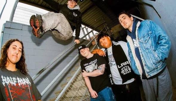 A group of five young men posing on a staircase, with one member performing a jump. They are dressed in casual streetwear, including hoodies and denim jackets.