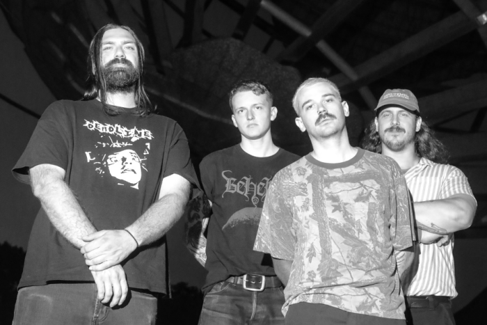 A group of four men posing together, dressed in casual clothing with dark and graphic t-shirts, in a partially darkened outdoor setting.