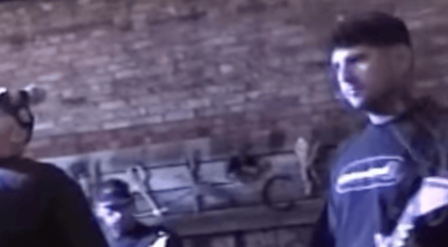 Two individuals standing in a dimly lit room with a brick wall in the background, one wearing headphones and the other holding an object.