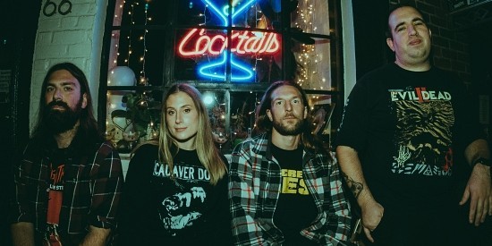 A group of four individuals posed in front of a neon 'Cocktails' sign, showcasing a vibrant nightlife atmosphere. The men have long hair and beards, while the woman in the center wears a casual shirt, with each person dressed in casual, graphic t-shirts and flannel shirts.