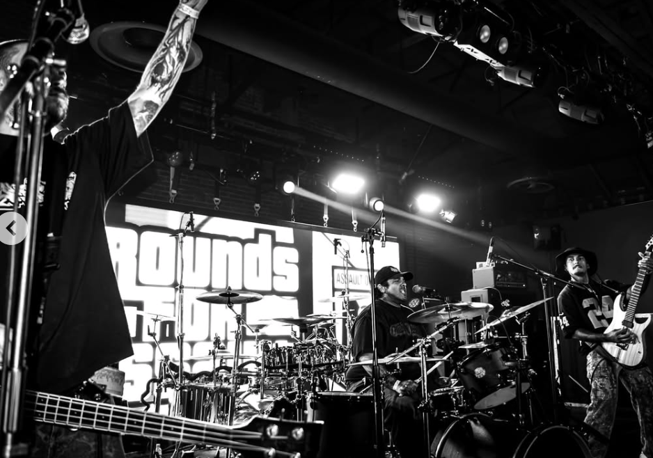 A black and white image of a live band performing on stage. The guitarist raises his hand while the drummer plays, and another musician is in the background. Bright stage lights and a large screen display are visible.