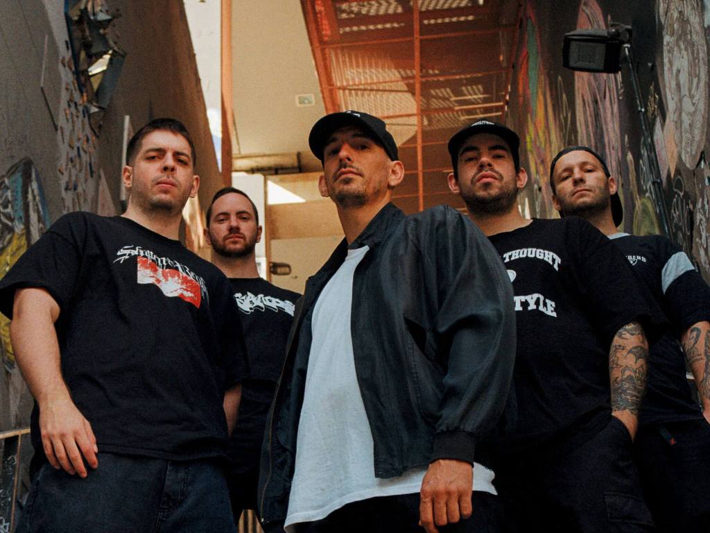 A group of five men posing confidently in an urban setting, wearing black t-shirts and relaxed clothing. They stand in a narrow alleyway adorned with colorful graffiti.