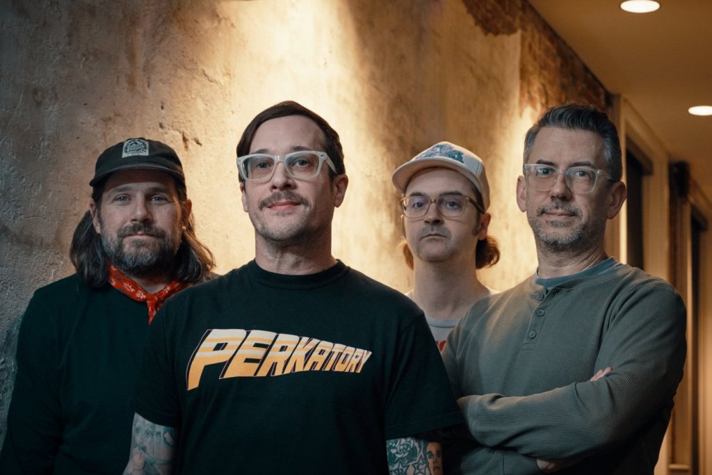 A group of four men posing together in a dimly lit indoor space, with a textured wall in the background. The man in the foreground is wearing a black 'PERKATORY' t-shirt, while the others have casual clothing. They are looking directly at the camera.