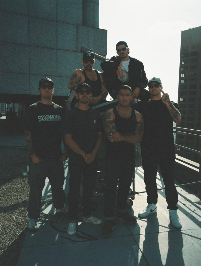 Group of six men with tattoos standing together on a rooftop against a city skyline, some wearing sunglasses and casual clothing.