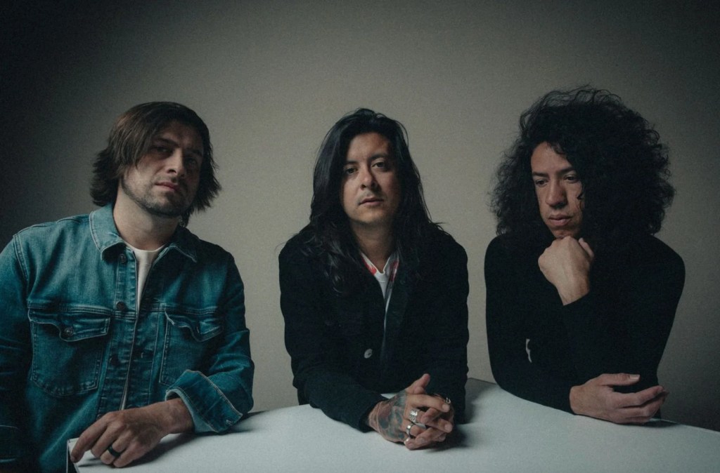 Three musicians posing together in a softly lit studio, with two wearing casual attire and one in black clothing, all seated at a table.
