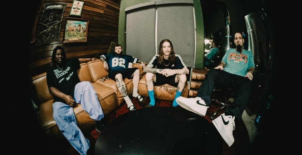 Four individuals seated on a brown leather couch in a casual indoor setting, with wooden walls and a round black table in front of them.