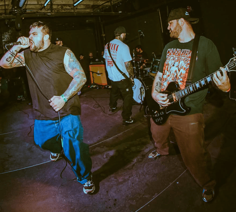 A hardcore band performing on stage, with a vocalist passionately singing into a microphone, a guitarist playing an electric guitar on the right, and a drummer visible in the background.