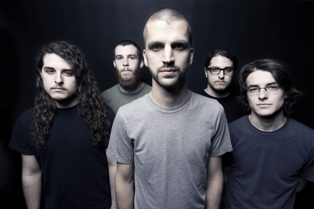 A group of five men standing close together, with varying hairstyles and facial hair, dressed in casual t-shirts. They are facing the camera against a dark background.